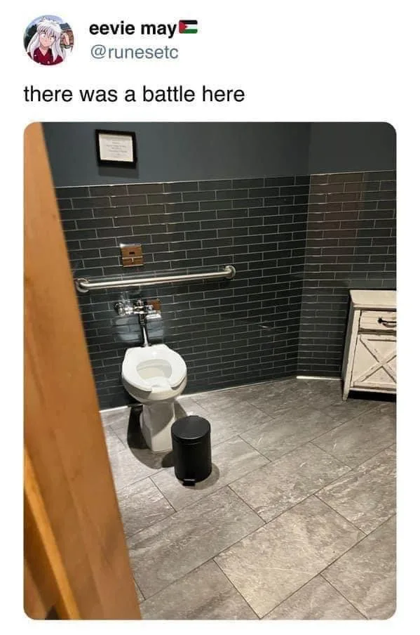 A photo of a clean, modern bathroom stall with dark gray tiled walls and floor, featuring a white toilet, a black trash can…