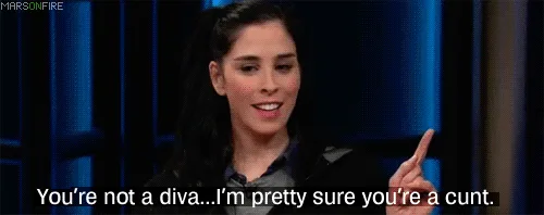 The image shows a woman with dark hair in a ponytail, wearing a dark blazer, speaking on a talk show set with blue vertical…