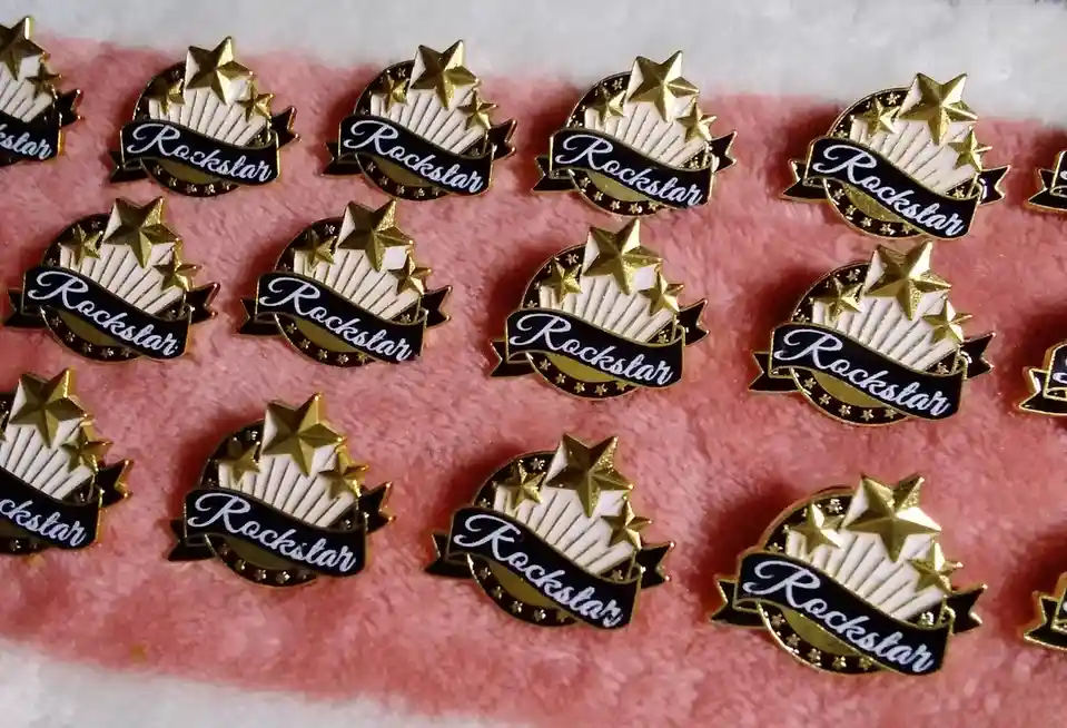 Multiple identical enamel pins featuring gold stars and a black banner with the word 'Rockstar' in white cursive text are…