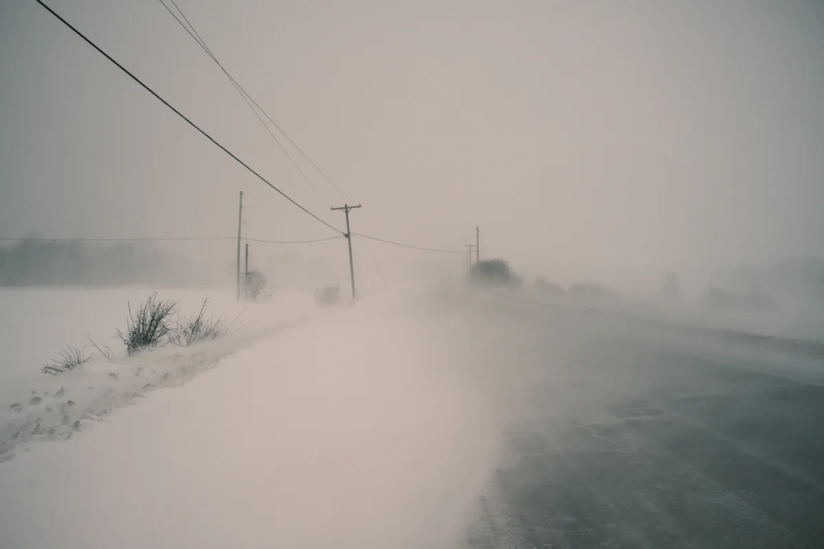 A desolate, snow-covered rural road stretches into a blizzard, with utility poles and wires visible against a gray, obscured…