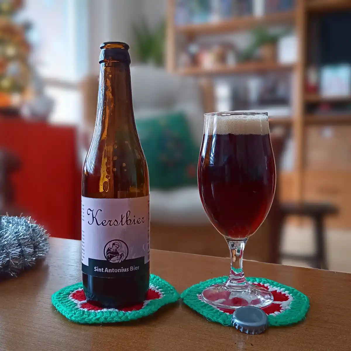 A brown beer bottle labeled 'Kerstbier Sint Antonius Bier' sits next to a glass of dark red beer on a wooden table, both…