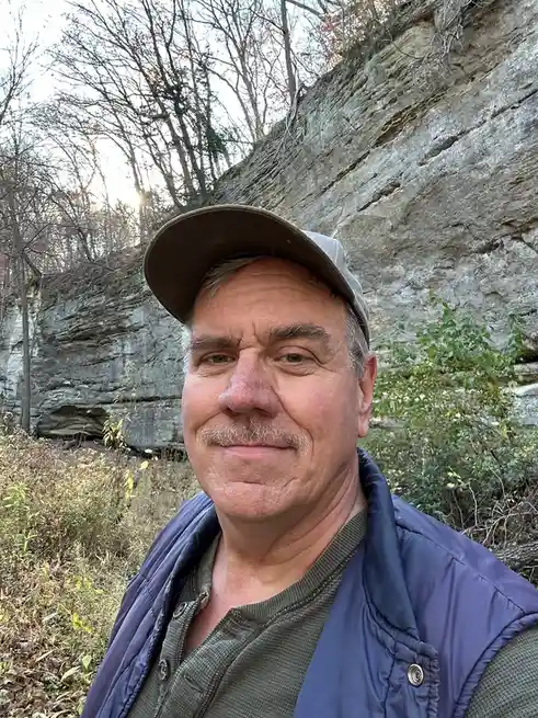 A middle-aged man with a mustache and a baseball cap takes a selfie in front of a large rock cliff with bare trees in the…