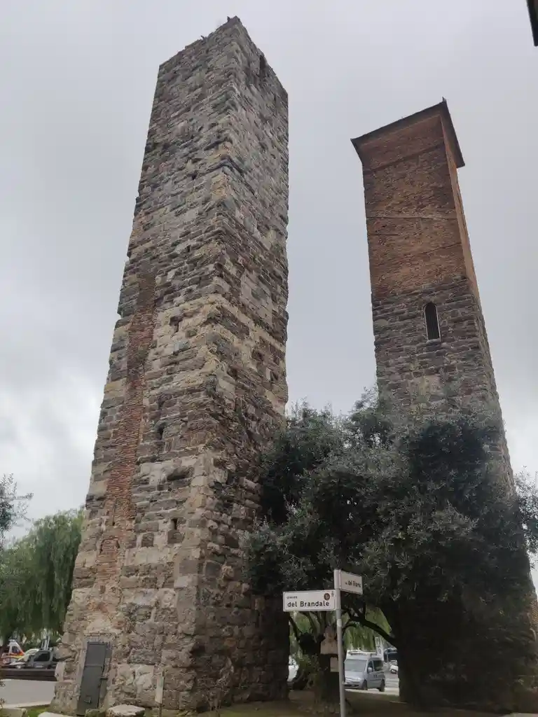Two tall, ancient stone towers stand side by side under an overcast sky, with a street sign reading 'piazza del Brandale'…