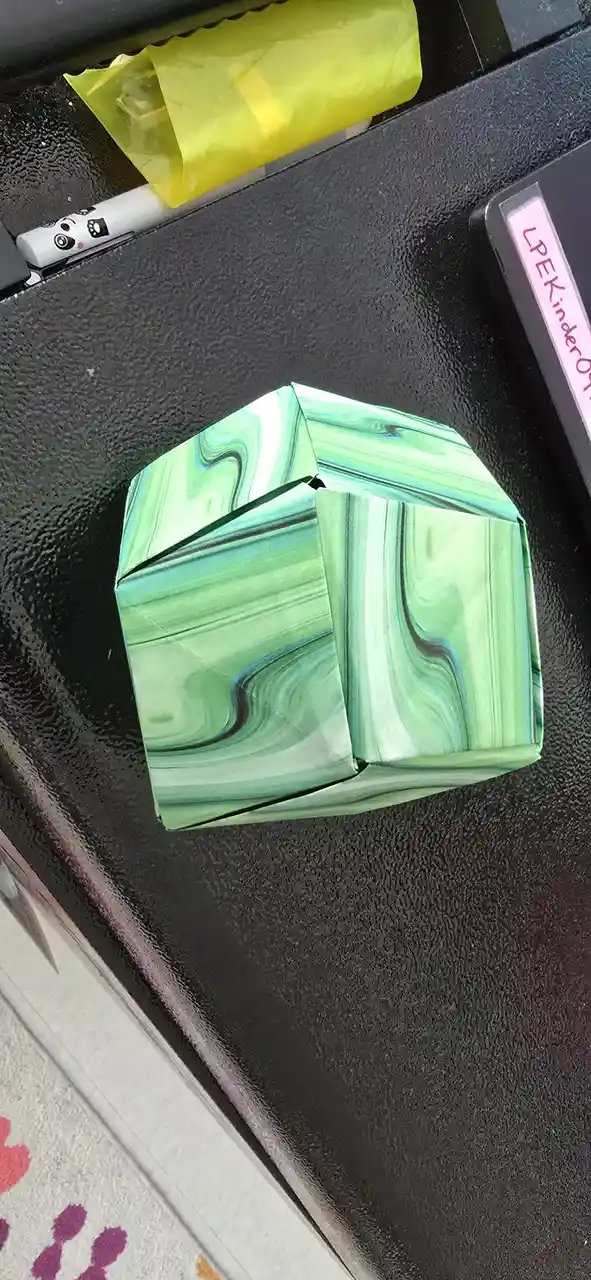 A handmade, green marbled paper cube sits on a black textured surface, with a yellow plastic bag and a white marker visible…