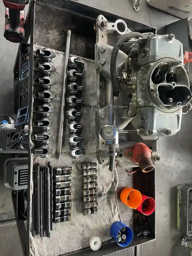 An overhead shot of a workbench displaying a disassembled carburetor next to an organized array of small metal parts…