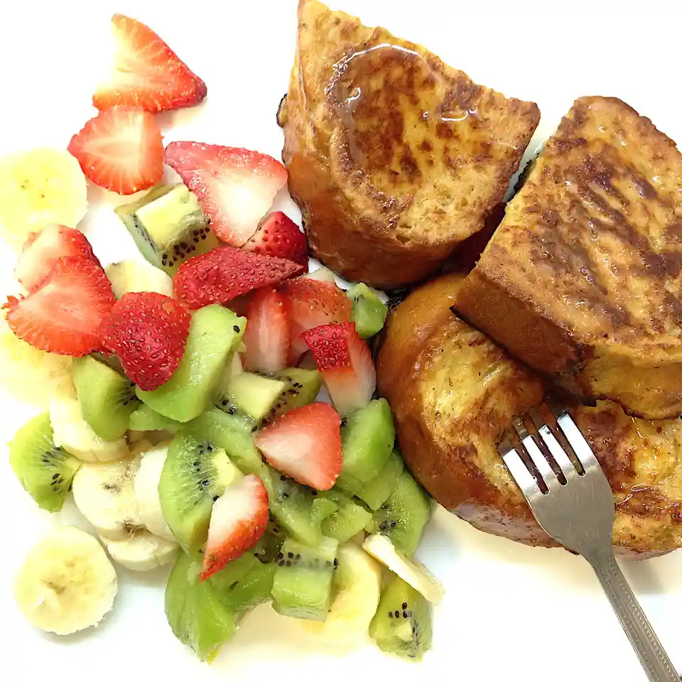 A plate of French toast drizzled with syrup sits next to a colorful fruit salad of sliced strawberries, kiwi, and banana. A…