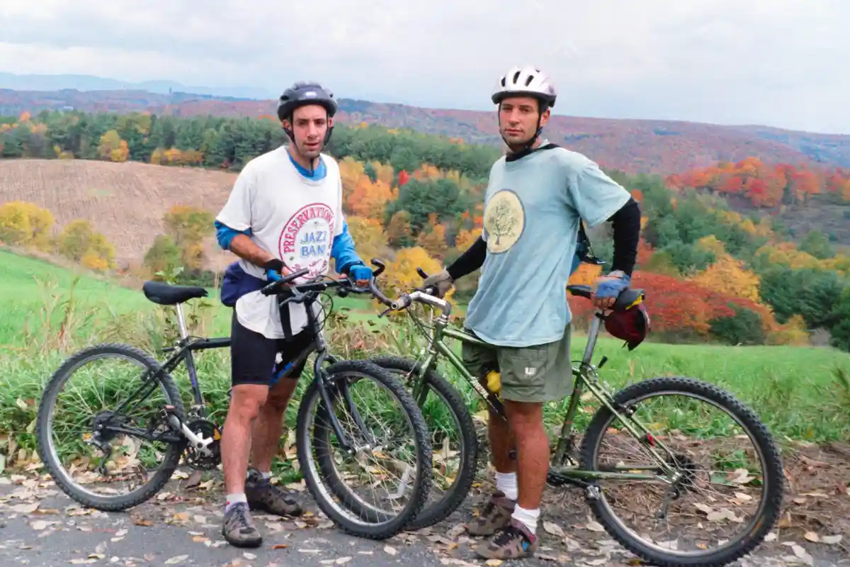 Two men in cycling gear stand beside their mountain bikes on a gravel path with autumn foliage in the background. The man on…
