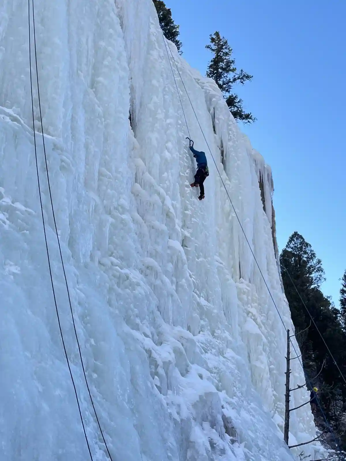 A person in winter gear is ice climbing up a massive frozen waterfall under a clear blue sky, secured by ropes. The climber…