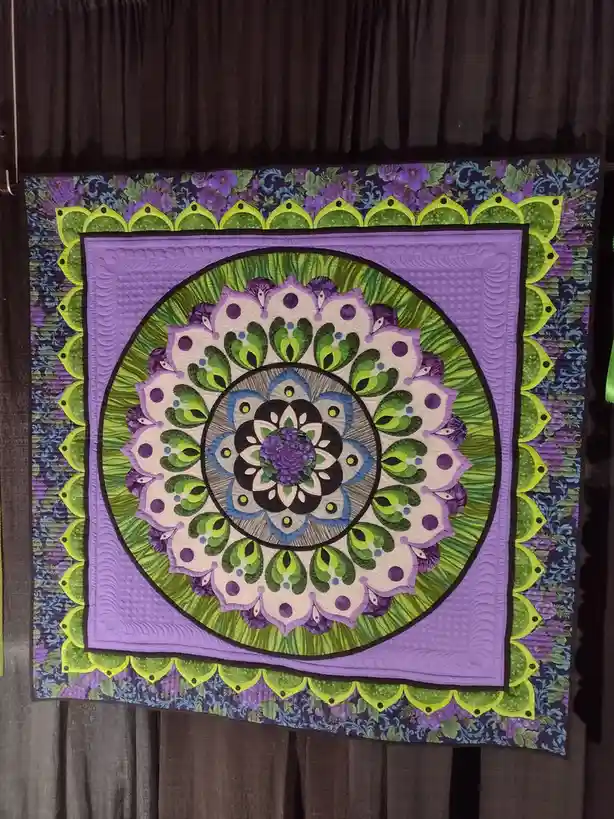 A colorful, intricately designed quilt with a symmetrical mandala pattern hangs against a dark curtain. The quilt features…