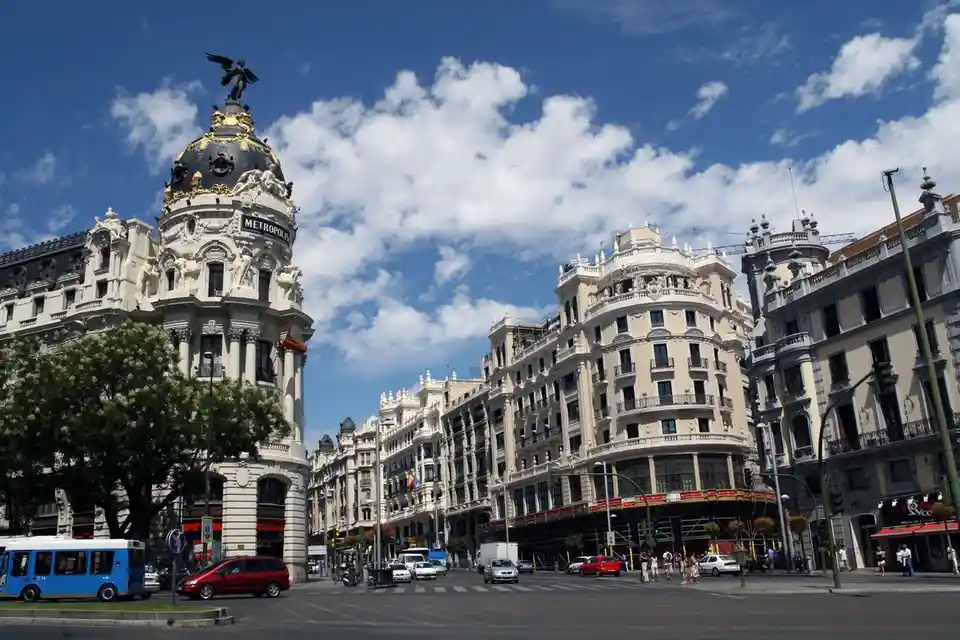 A wide-angle photograph of a sunny urban street corner featuring ornate, historic European architecture under a blue sky…