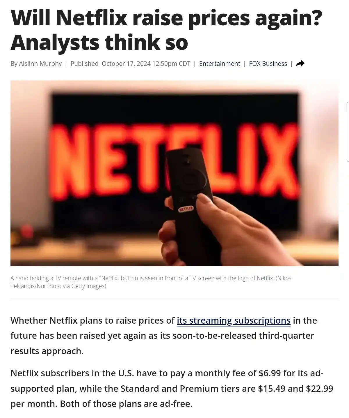 A hand holds a black Netflix-branded TV remote in front of a TV screen displaying the red Netflix logo. The image is from a…