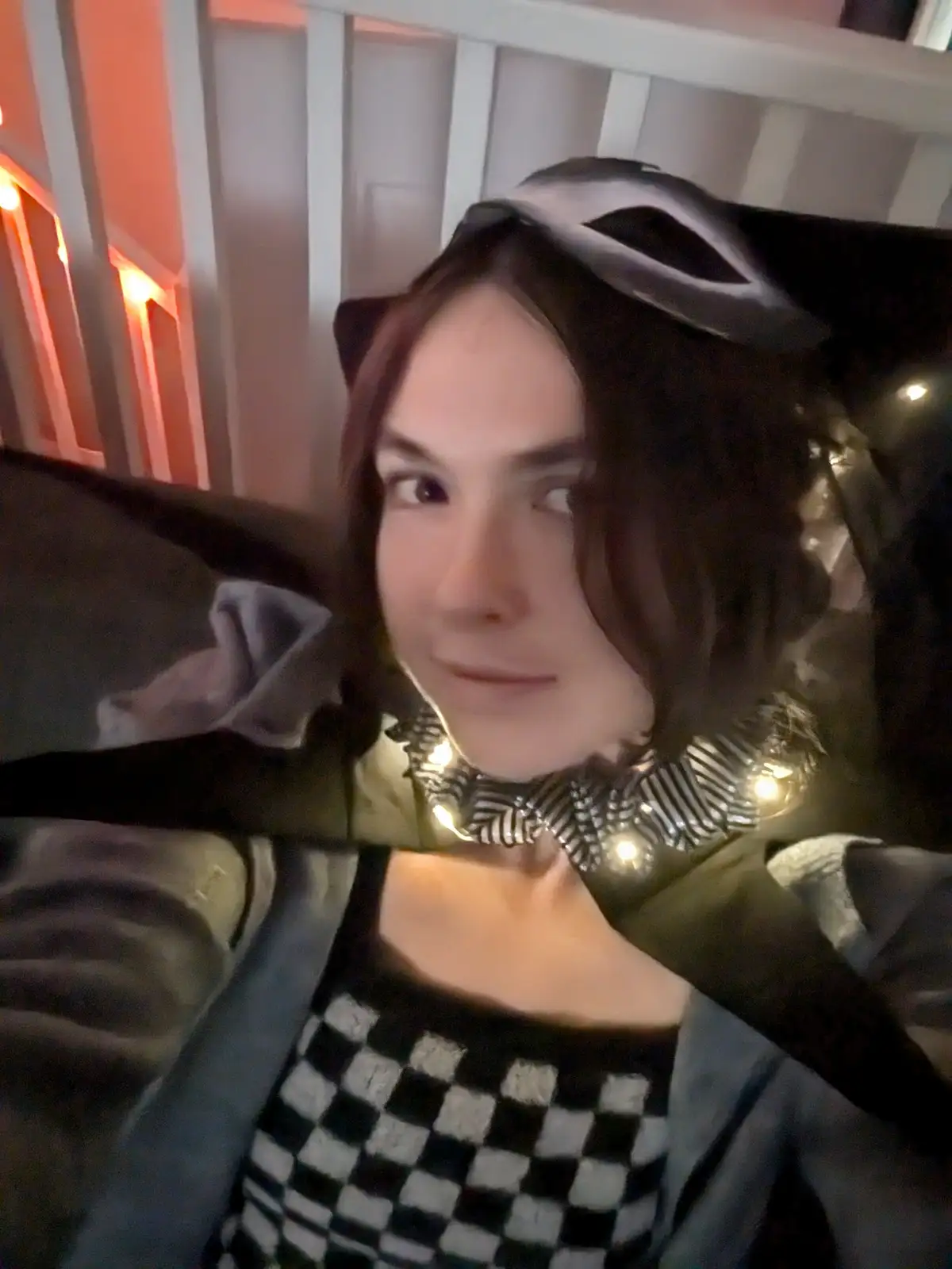 A person with dark hair wears a black cat ear headband and a black-and-white checkered top under a gray jacket. They have a…