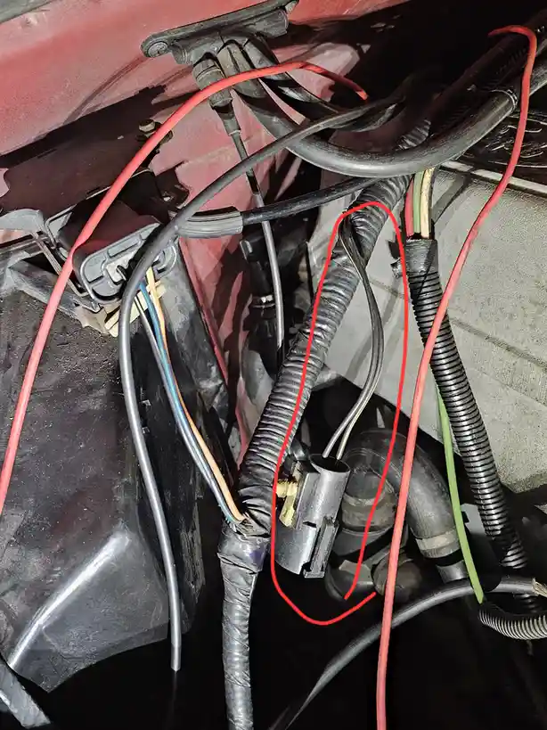 a close-up photo of a car's engine bay wiring harness, showing a single electrical connector circled in red amidst a tangle…