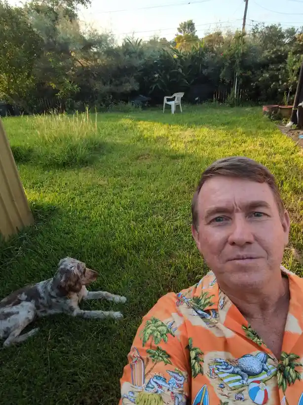 A man in a bright orange Hawaiian shirt with alligator and palm tree prints takes a selfie in a grassy backyard, looking…