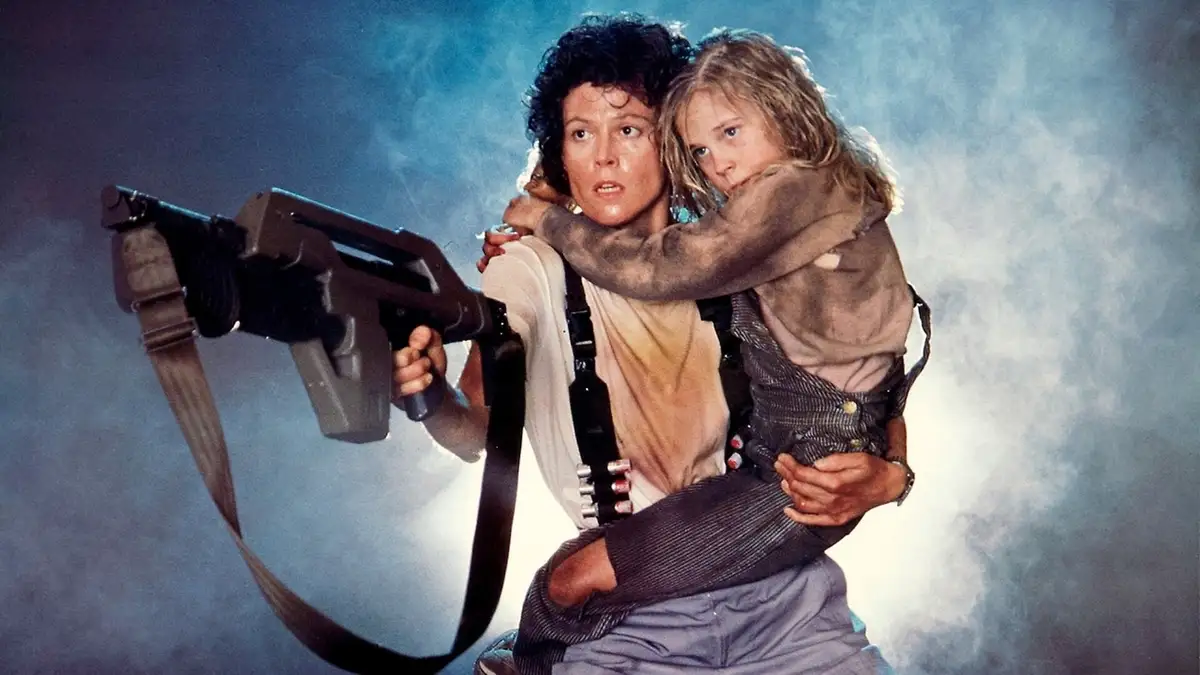 A woman with curly dark hair, holding a large sci-fi weapon, carries a young girl in a dirty outfit. Both look tense against…