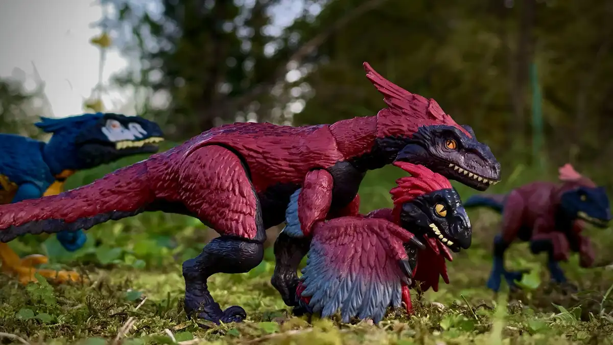A red and black dinosaur toy figurine is posed on mossy ground, appearing to be attacking or holding down a smaller…