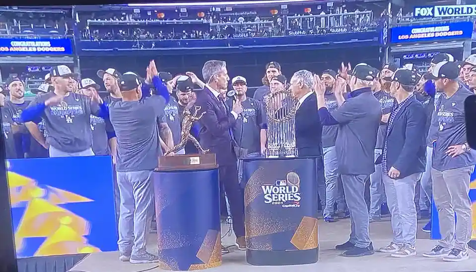A group of Los Angeles Dodgers players and staff in gray 'World Series Champions' t-shirts celebrate on stage with two…