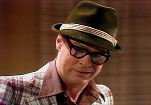a man wearing a green fedora with a light-colored band and thick black-rimmed glasses, looking slightly smug or dismissive…