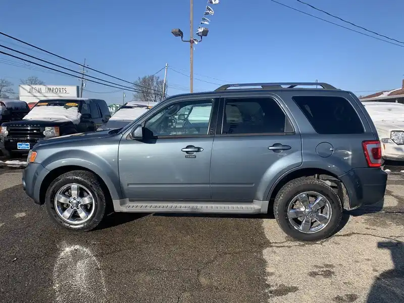 A side profile photo of a gray Ford Escape SUV parked in a car lot under a clear blue sky. The vehicle has silver roof…