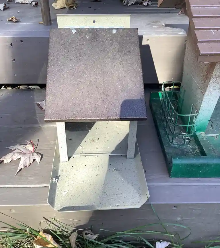A close-up photo shows a small, beige plastic outdoor table with a speckled brown top, sitting on a wooden deck next to a…