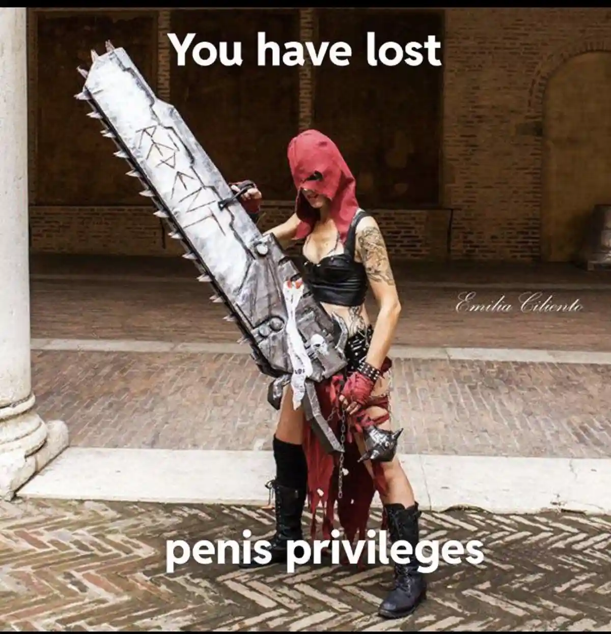A person in a red hooded costume holding a large, spiked chainsaw-like weapon stands in a courtyard. White text overlaid at…
