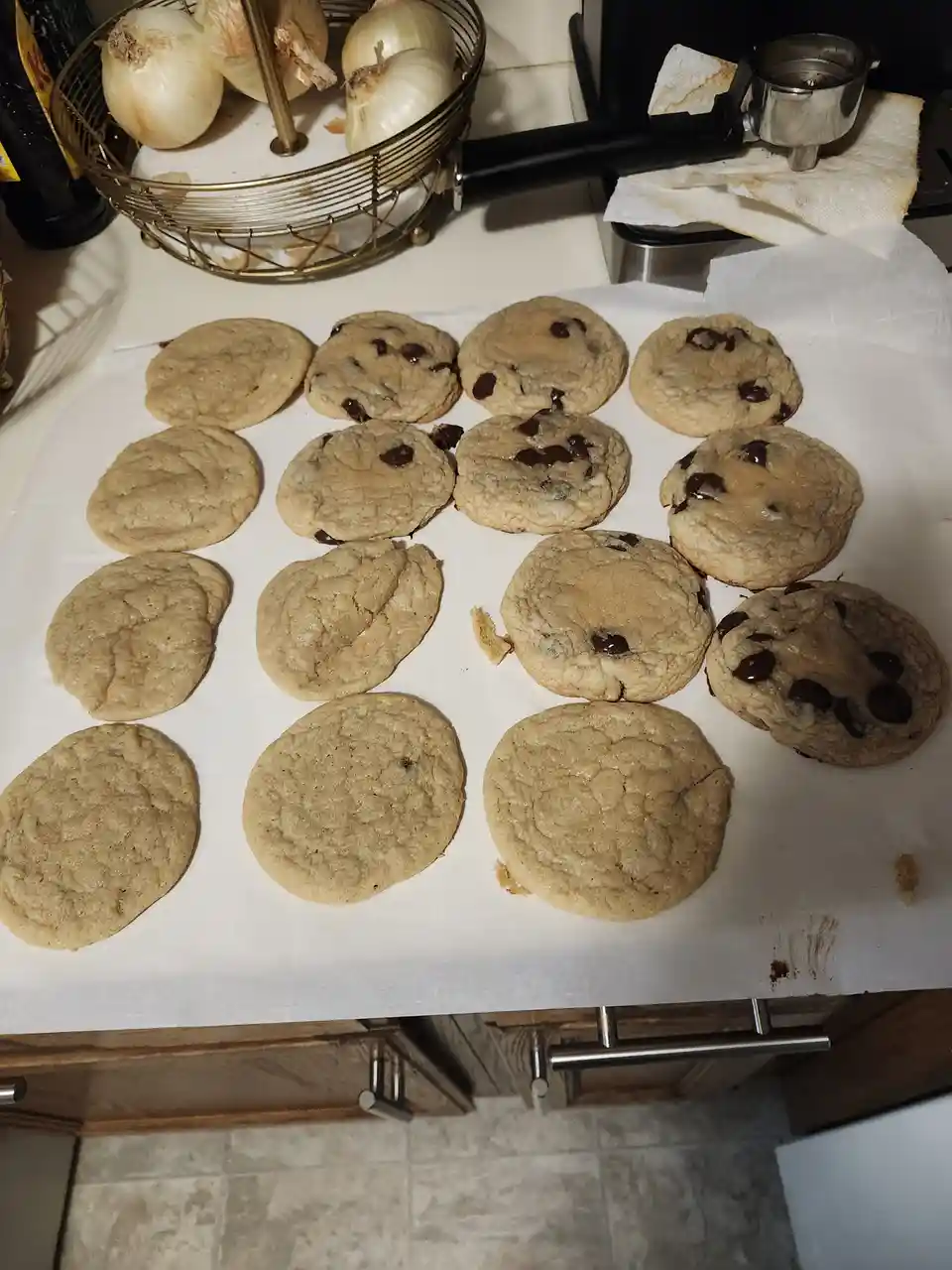 A kitchen counter displays a batch of freshly baked chocolate chip cookies on parchment paper, with some cookies appearing…
