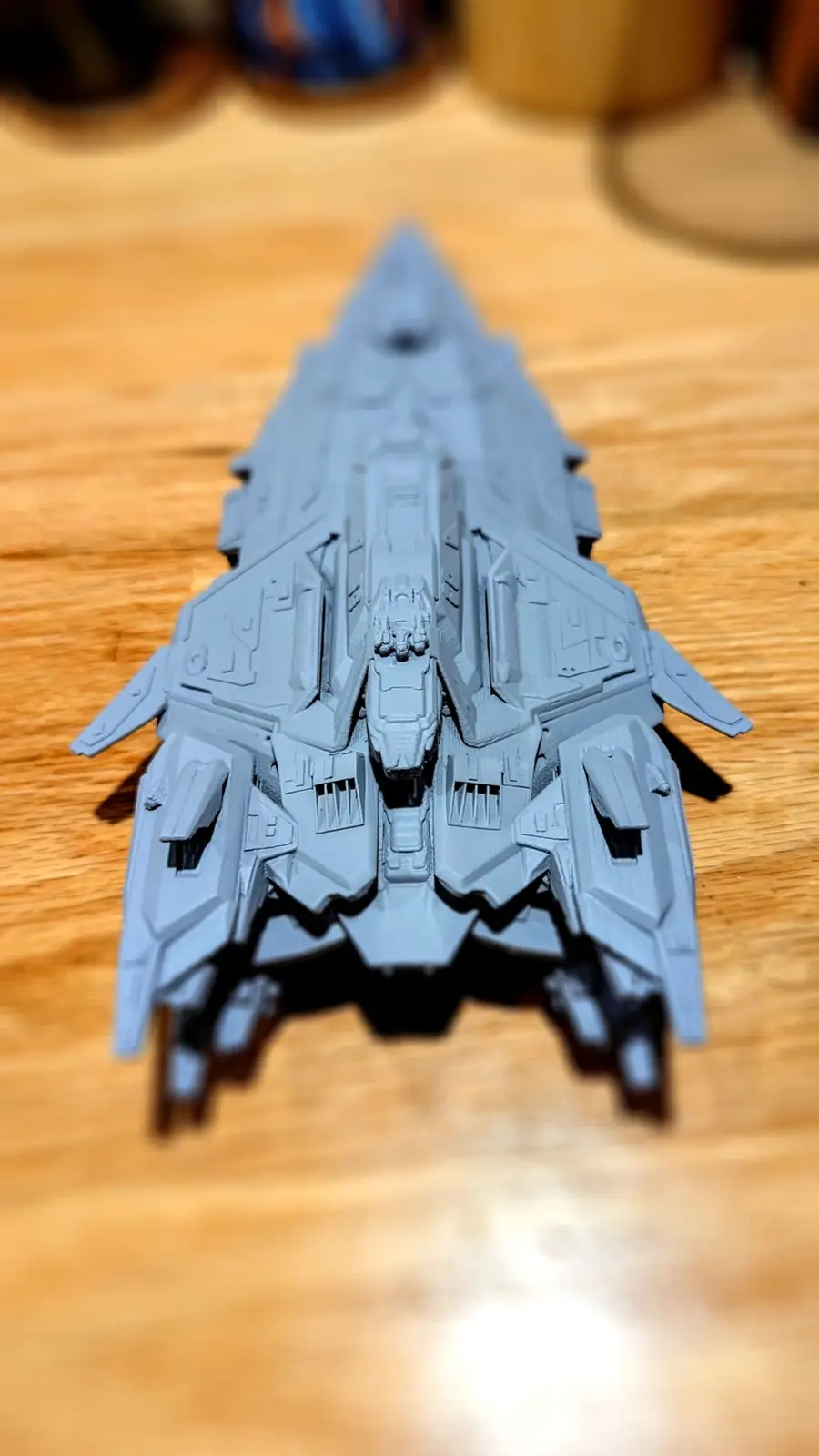 A gray, intricately detailed model of a futuristic spaceship sits on a wooden table, viewed from the front. The overlaid…