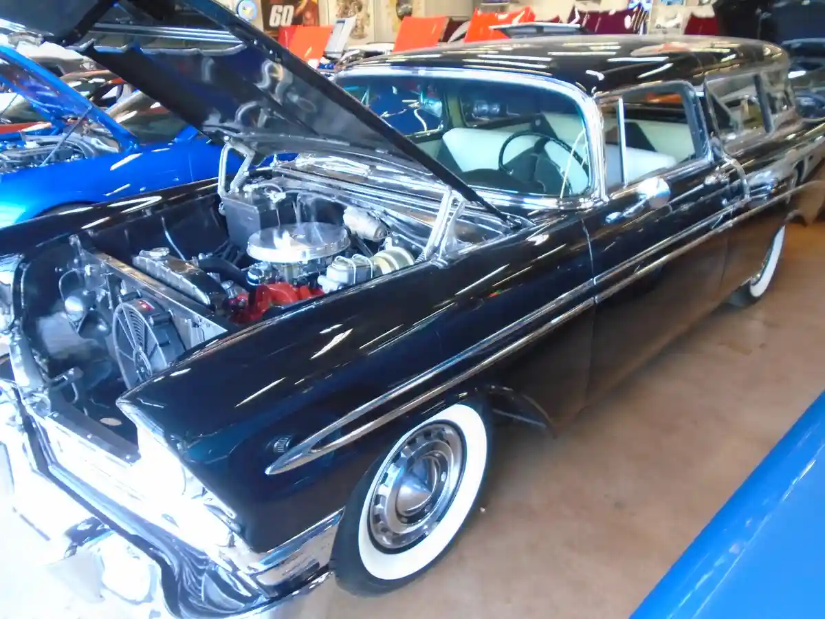 A glossy black classic American car with its hood open, revealing a shiny engine, is parked indoors among other vintage…
