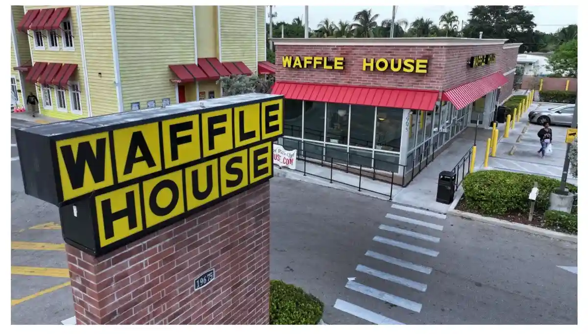 An exterior photo of a Waffle House restaurant with its iconic yellow and black sign on a brick pillar in the foreground…