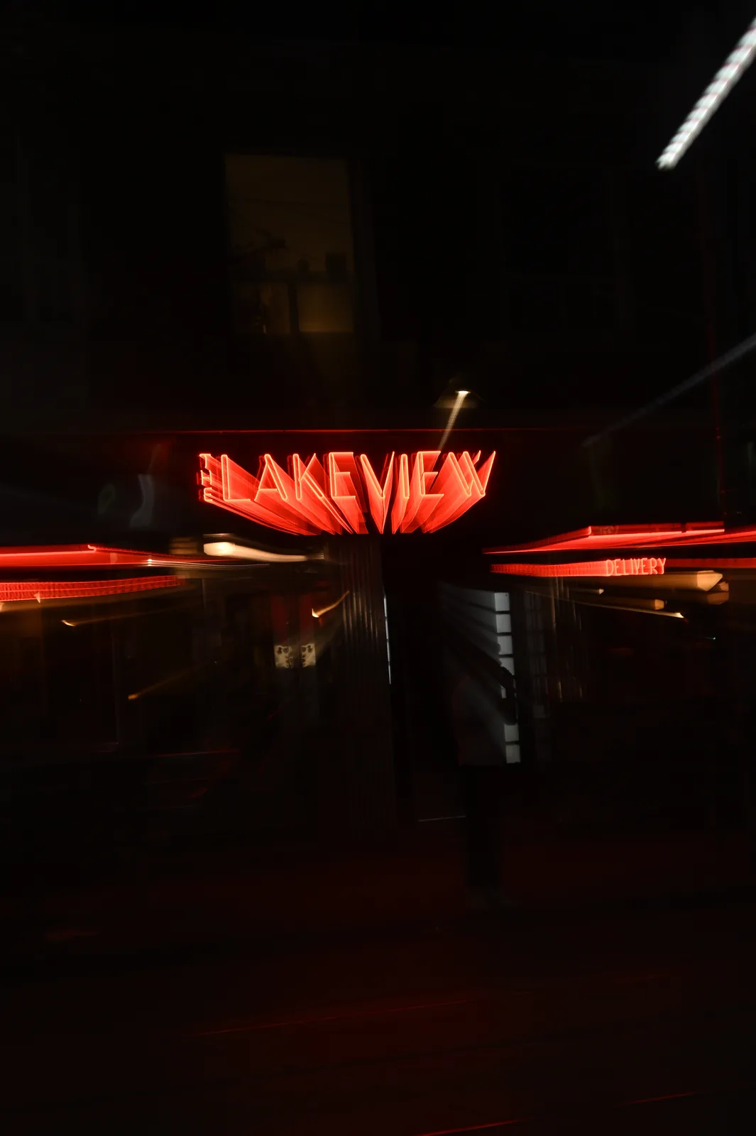 A long-exposure night photo shows a blurred, red neon sign reading 'LAKEVIEW' above a storefront with a smaller 'DELIVERY'…