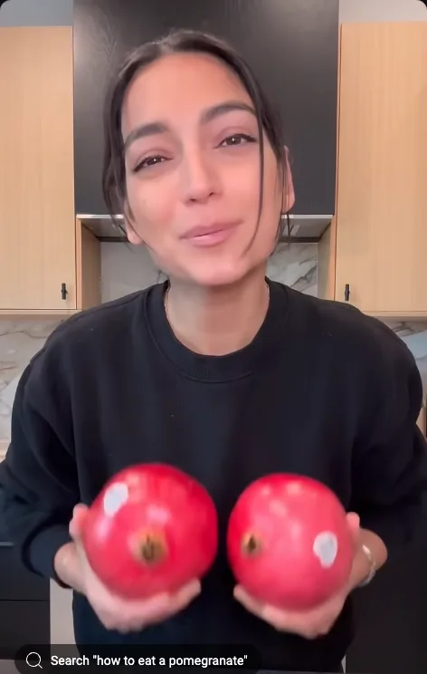 A woman in a black sweatshirt stands in a kitchen holding two pomegranates, each with a white sticker on it. She looks at…