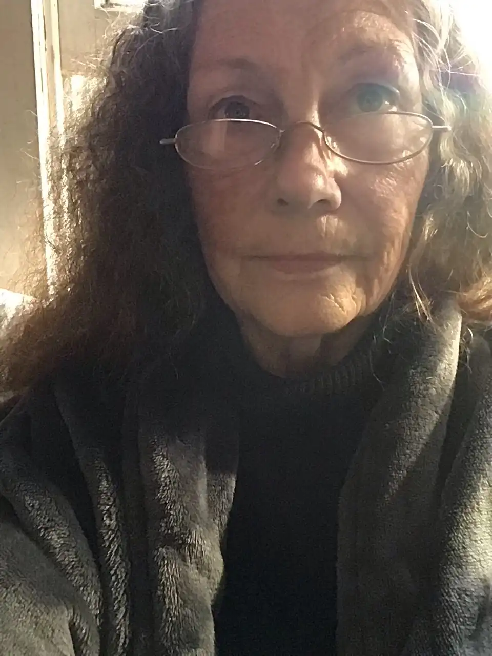 A close-up photo of an older woman with curly hair and glasses, wearing a dark turtleneck and a grey fleece robe, looking…