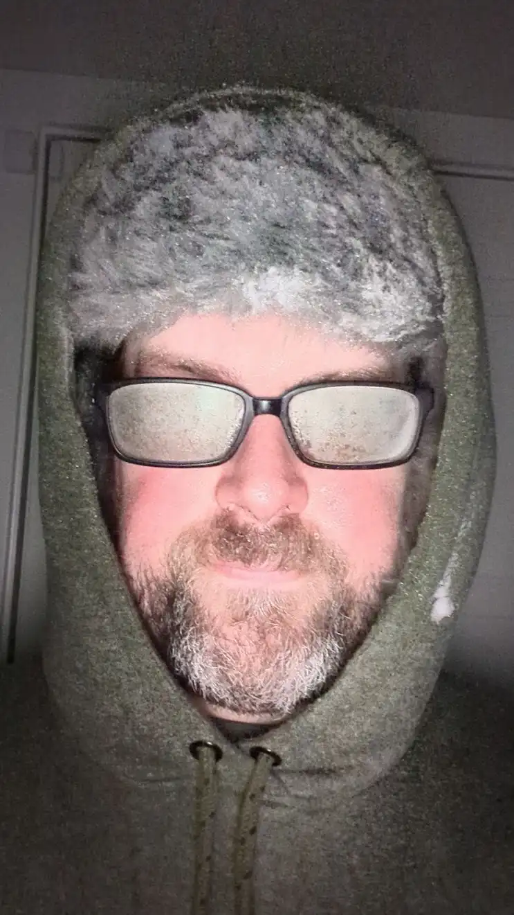A man wearing a green hooded sweatshirt with the hood up, covered in frost and ice crystals on the hood and glasses. His…