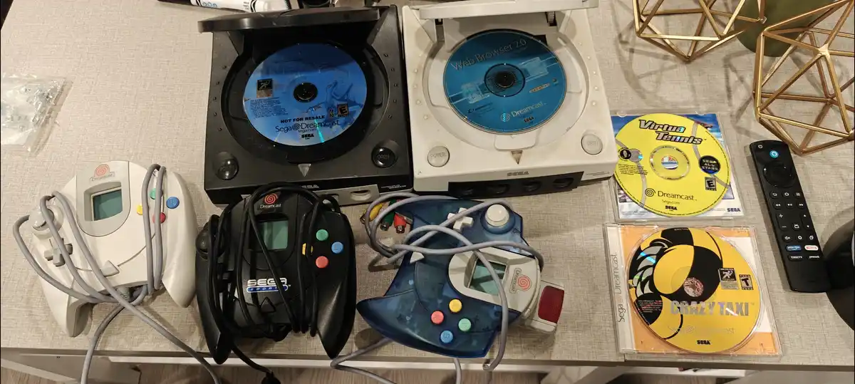 Two Sega Dreamcast consoles sit side-by-side on a table, one black and one white, both with open disc trays showing game…