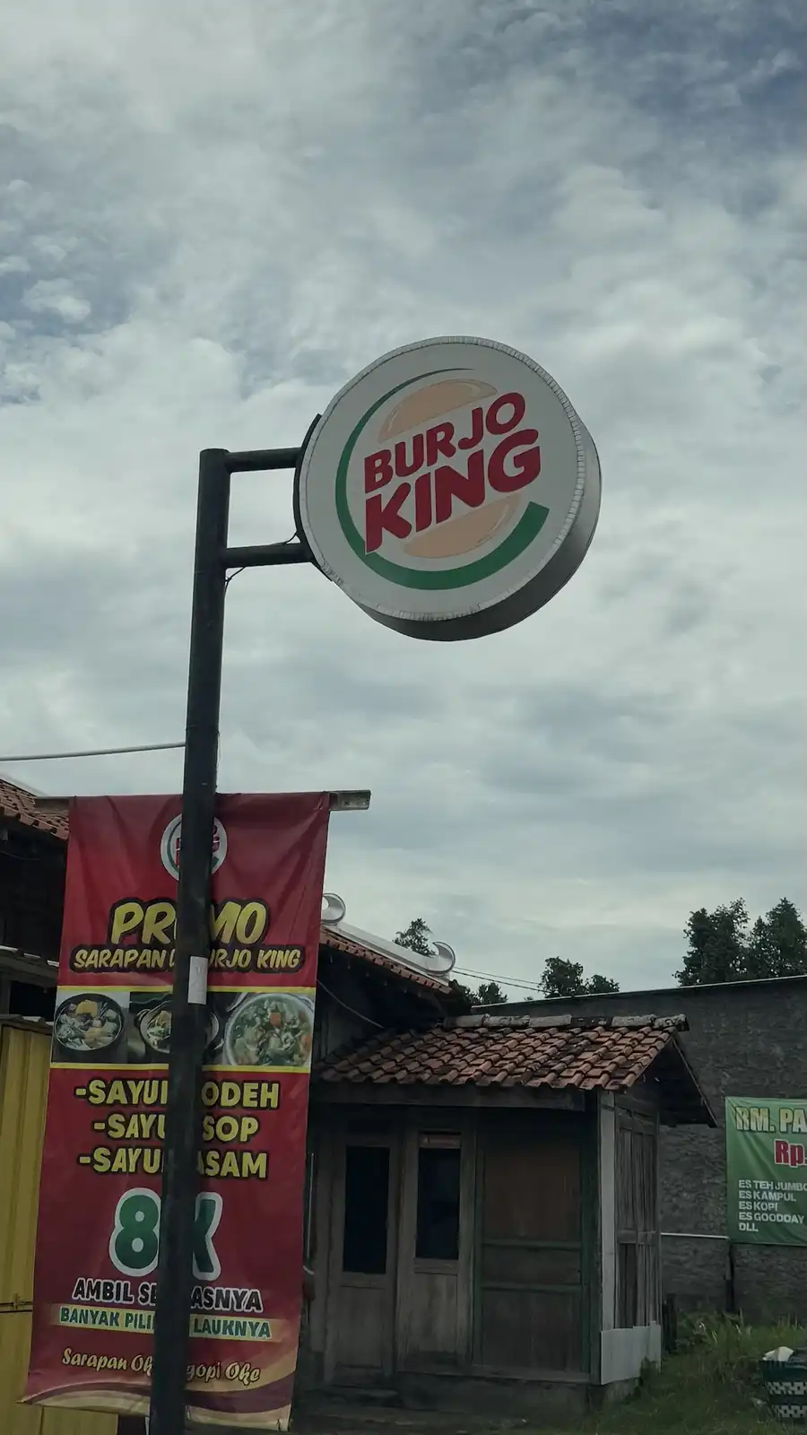 A circular sign for 'BURJO KING' with a burger-like logo hangs above a modest, rustic-looking eatery with a tiled roof. A…