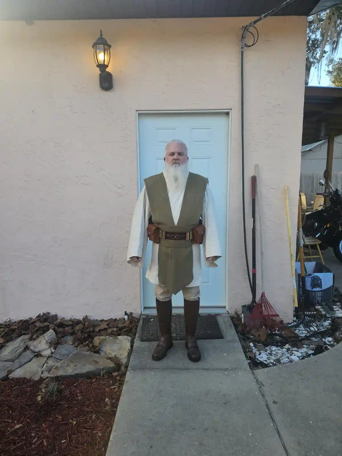 A man with a long white beard stands in front of a house door wearing a full Jedi Master costume from Star Wars, complete…