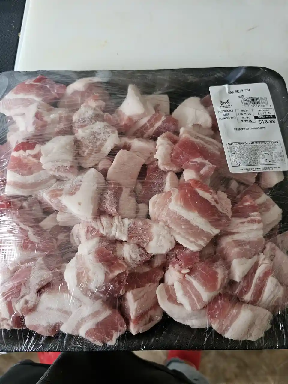 A close-up photo of a black plastic tray filled with raw, cubed pork belly, covered in plastic wrap, with a Member's Mark…