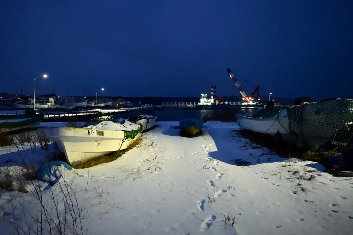 A snowy harbor at night with several small boats resting on the snow-covered ground, one prominently displaying the…