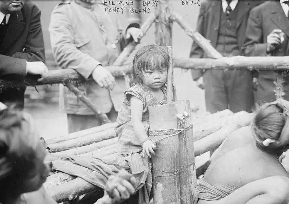 A vintage black-and-white photograph shows a young child with a solemn expression, standing beside a bamboo pole while…