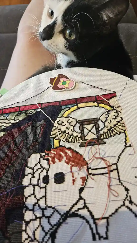 A black and white cat with wide, alert eyes looks down at a partially completed cross-stitch project featuring a pixelated…