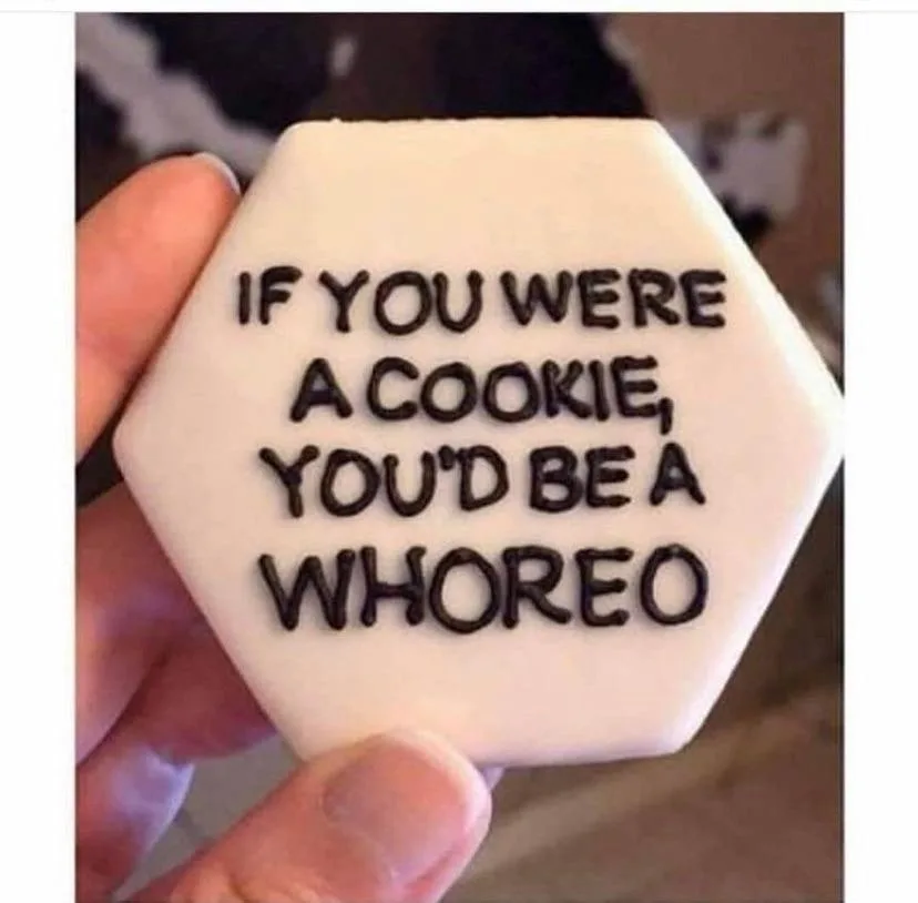 A hand holds a hexagonal white cookie with black icing text that reads 'IF YOU WERE A COOKIE, YOU'D BE A WHOREO'. The cookie…