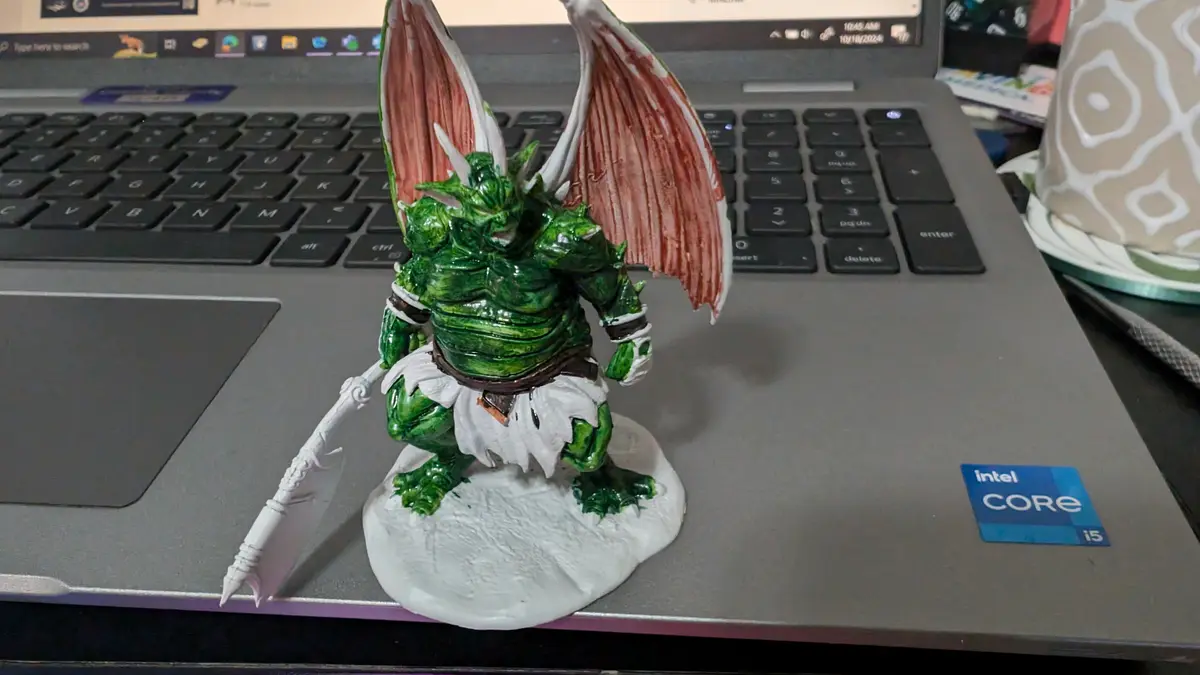 A green, muscular, winged fantasy creature figurine with red-striped wings and a white loincloth stands on a laptop…