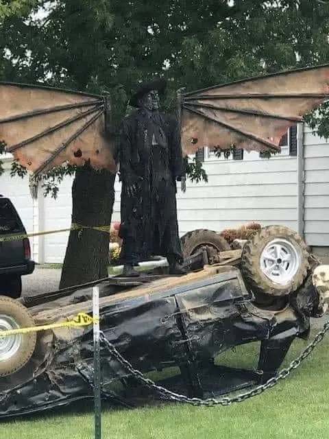 a large, black, winged demon statue stands atop a flipped-over black car, which is chained and cordoned off with yellow…