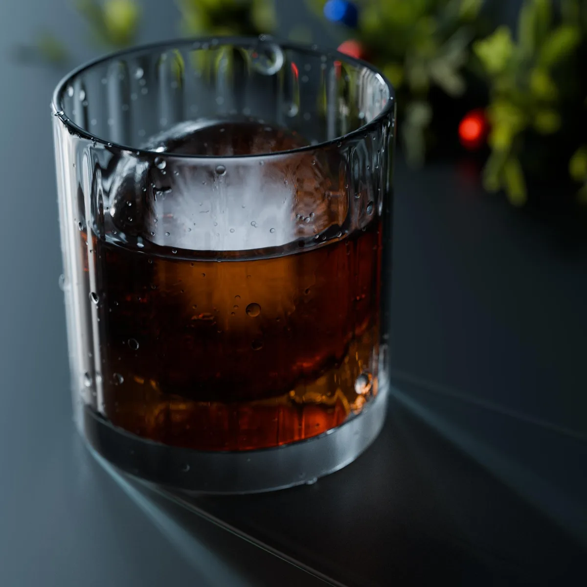 A close-up of a chilled rocks glass filled with amber-colored liquid and a large spherical ice cube, condensation beads on…
