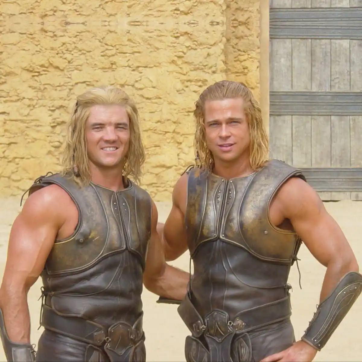 Two men in ancient warrior armor stand side by side against a textured yellow wall and wooden door. The man on the left has…