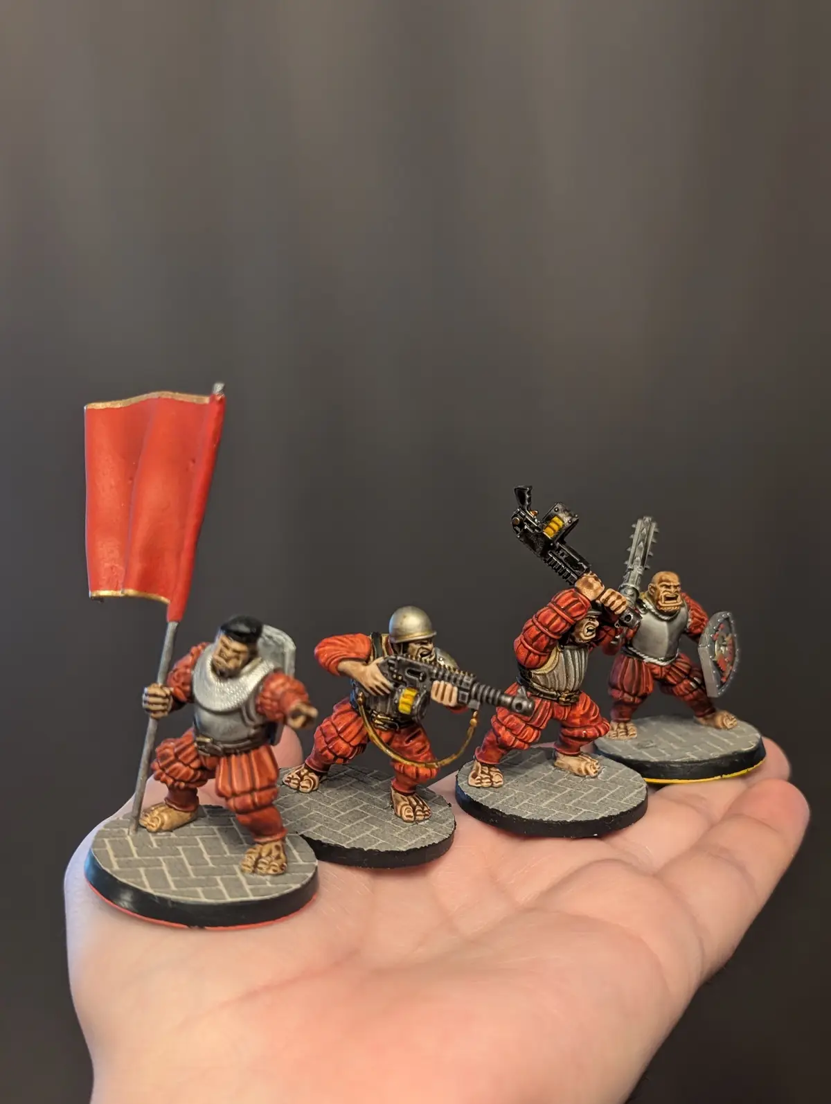 A person's open palm holds four small, painted fantasy miniatures of ork warriors in red and silver armor. One carries a red…