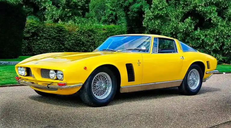 A bright yellow vintage sports car, likely a Maserati, is parked on a gravel driveway with lush green hedges in the…