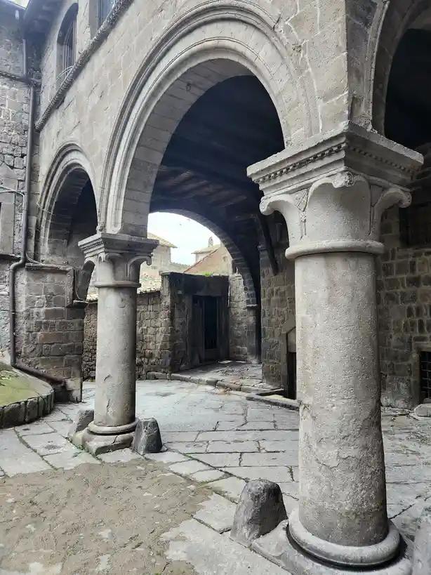 The image shows a stone courtyard with two large, ornate columns supporting arched walkways, typical of old European…