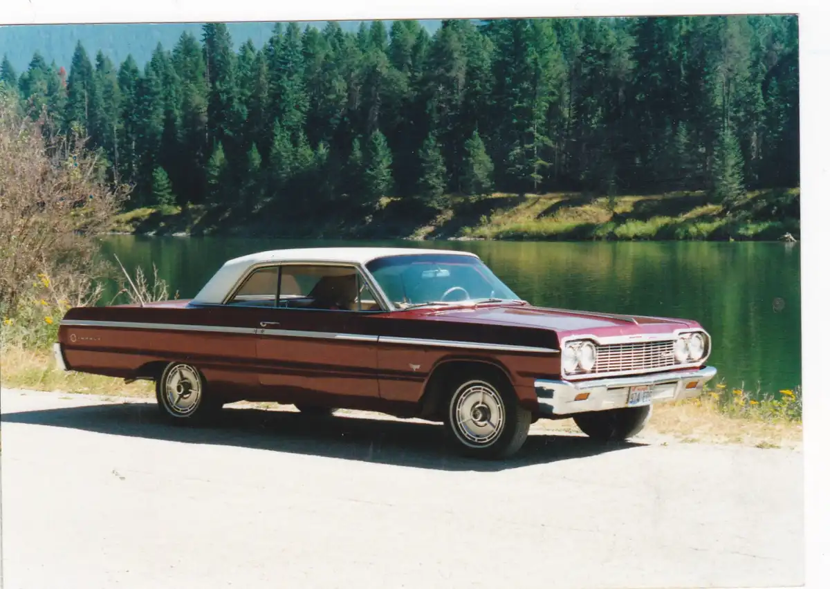 A vintage maroon and white two-tone car, likely a 1960s American sedan, is parked on a gravel road beside a calm green lake…
