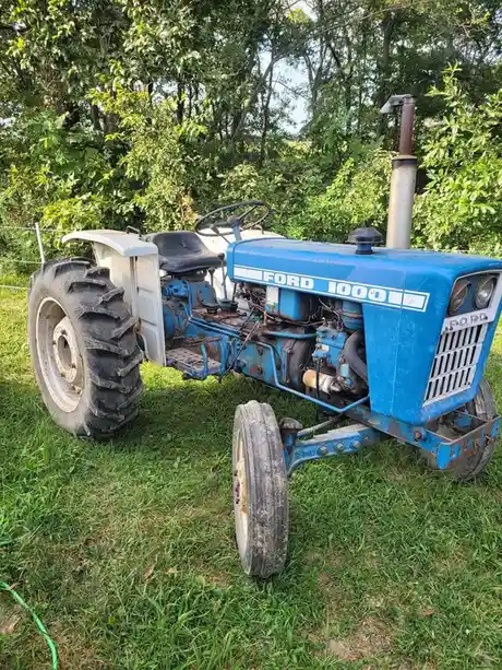 A blue and white Ford 1000 tractor sits on grass with trees in the background, showing visible wear and tear. The text 'ford…